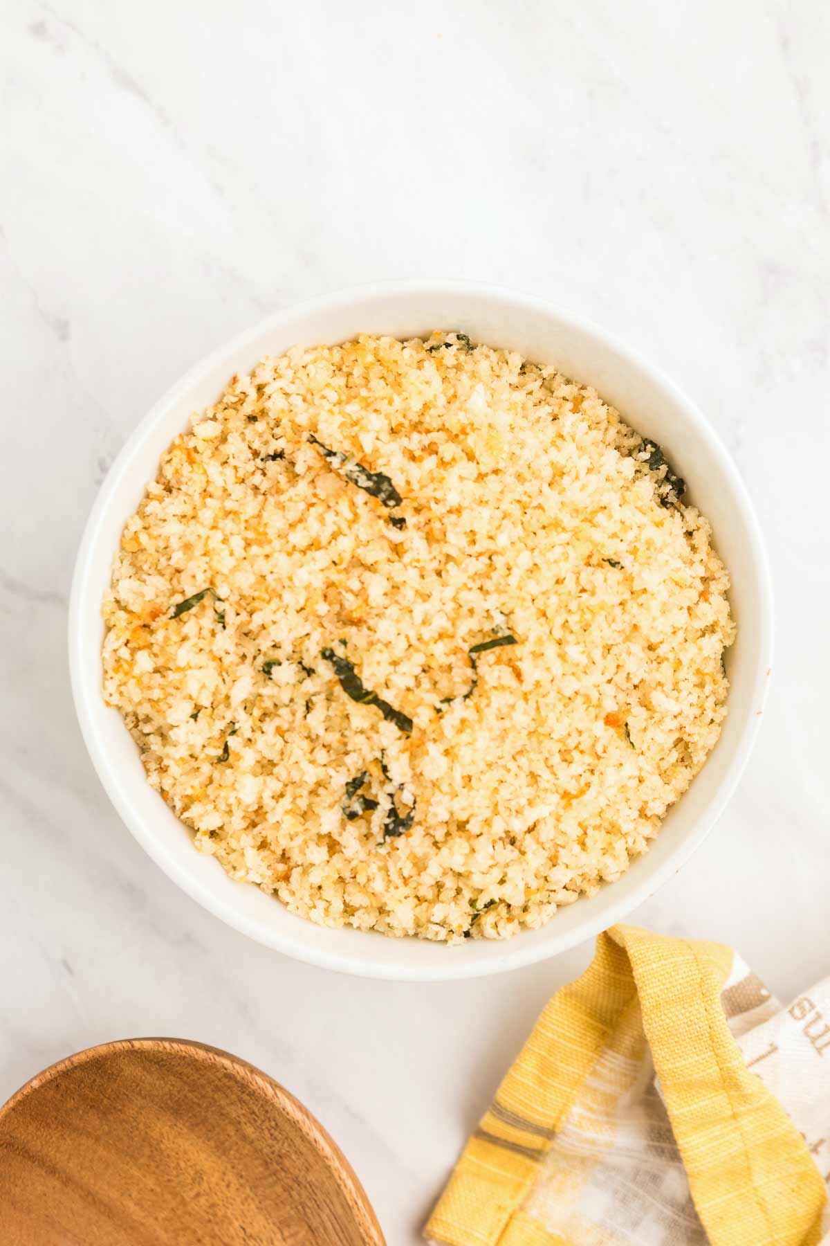 A bowl of Panko bread crumbs with herbs sits on a white marble surface. A yellow cloth and a wooden utensil are nearby.
