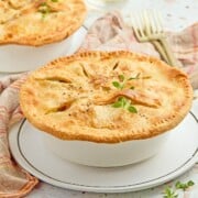 A golden-brown pot pie in a white dish on a plate, garnished with herbs, with a fork and napkin nearby.