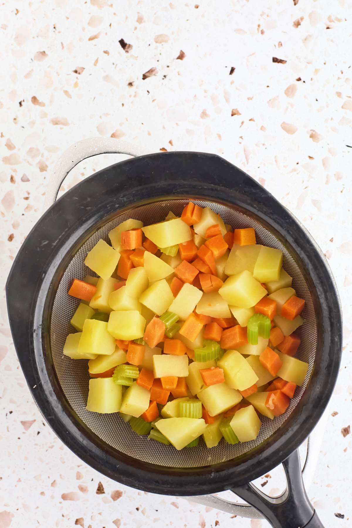 A strainer with diced boiled vegetables, including potatoes, carrots, and celery, on a speckled countertop.