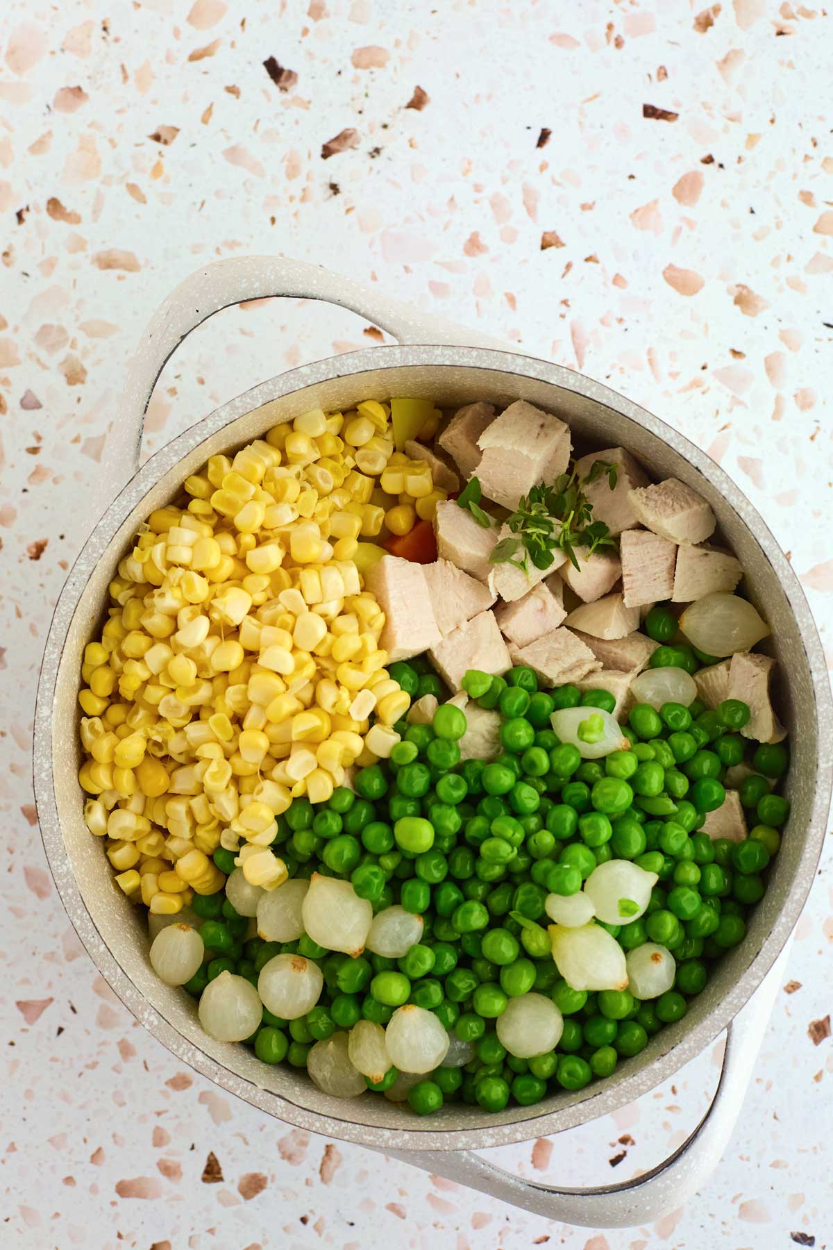 A pot filled with corn, peas, pearl onions, and diced meat on a terrazzo countertop.