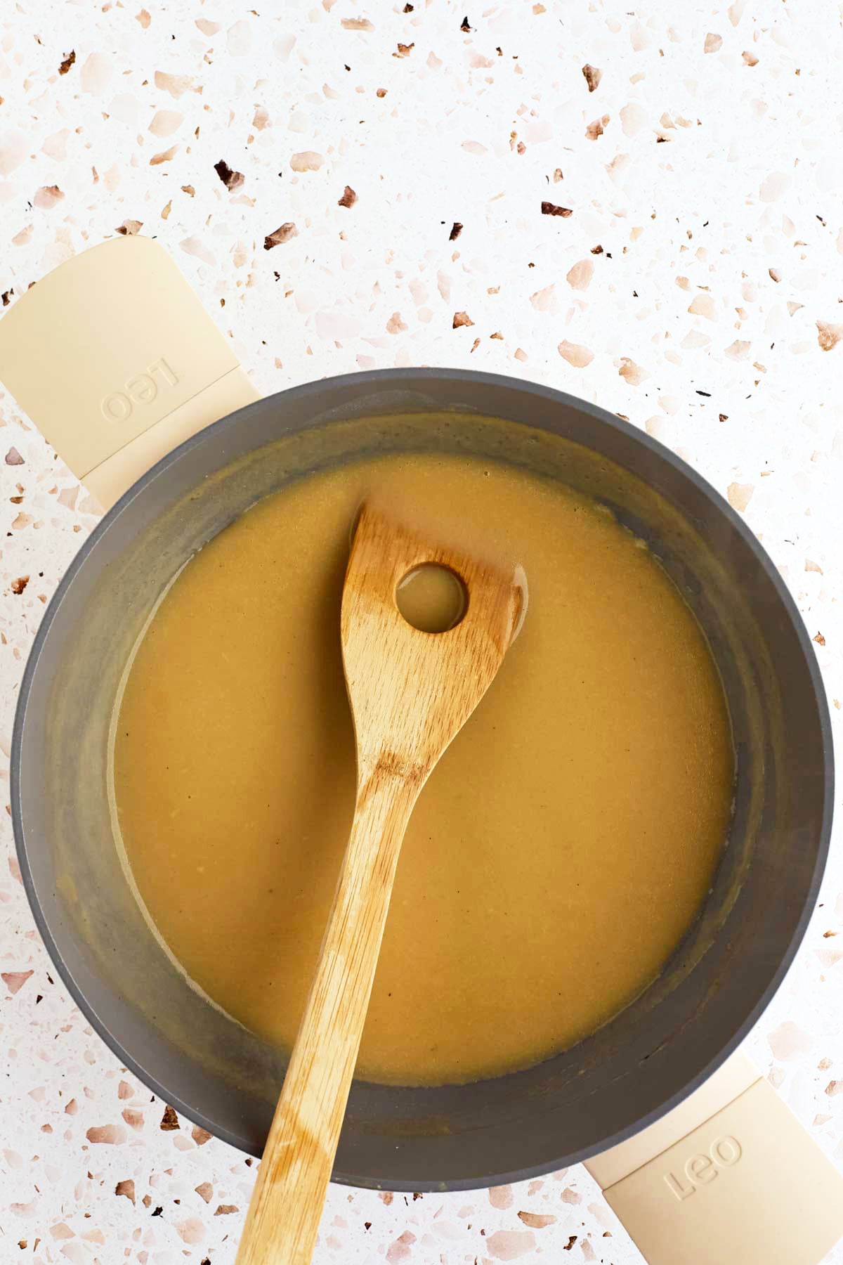 A wooden spoon rests in a pot of thick brown gravy on a speckled countertop.