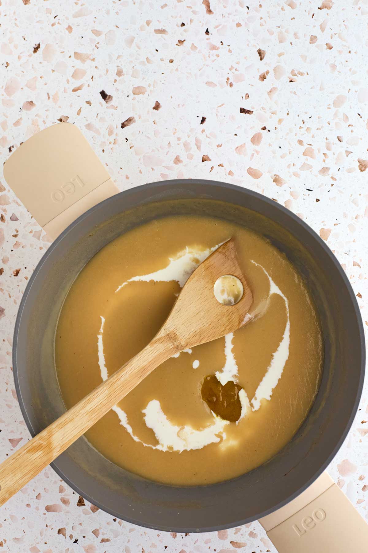 A pot of creamy soup with a wooden spoon and a swirl of cream on top, set on a speckled countertop.