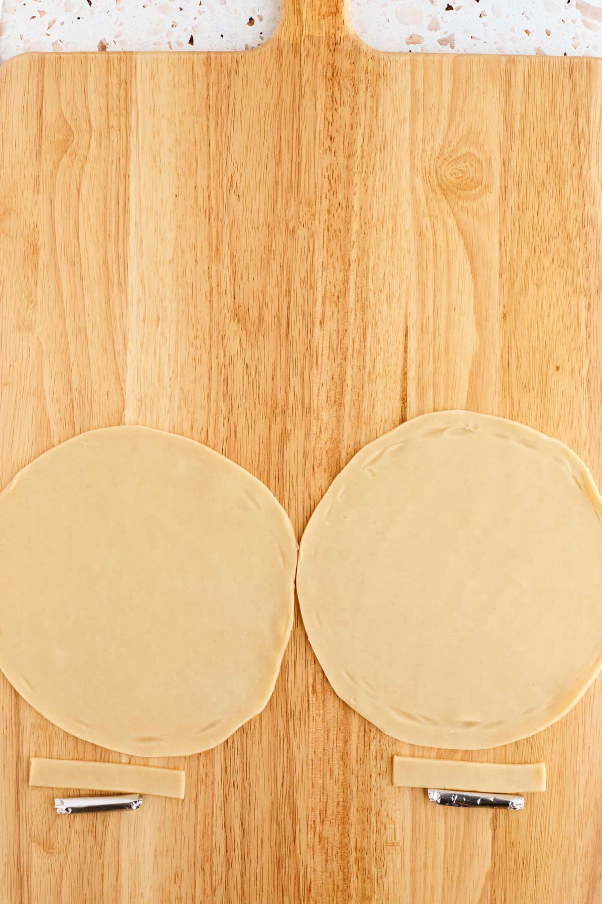 Two round dough circles and two rectangular dough strips on a wooden surface, with a crimping tool and a small rolling pin.