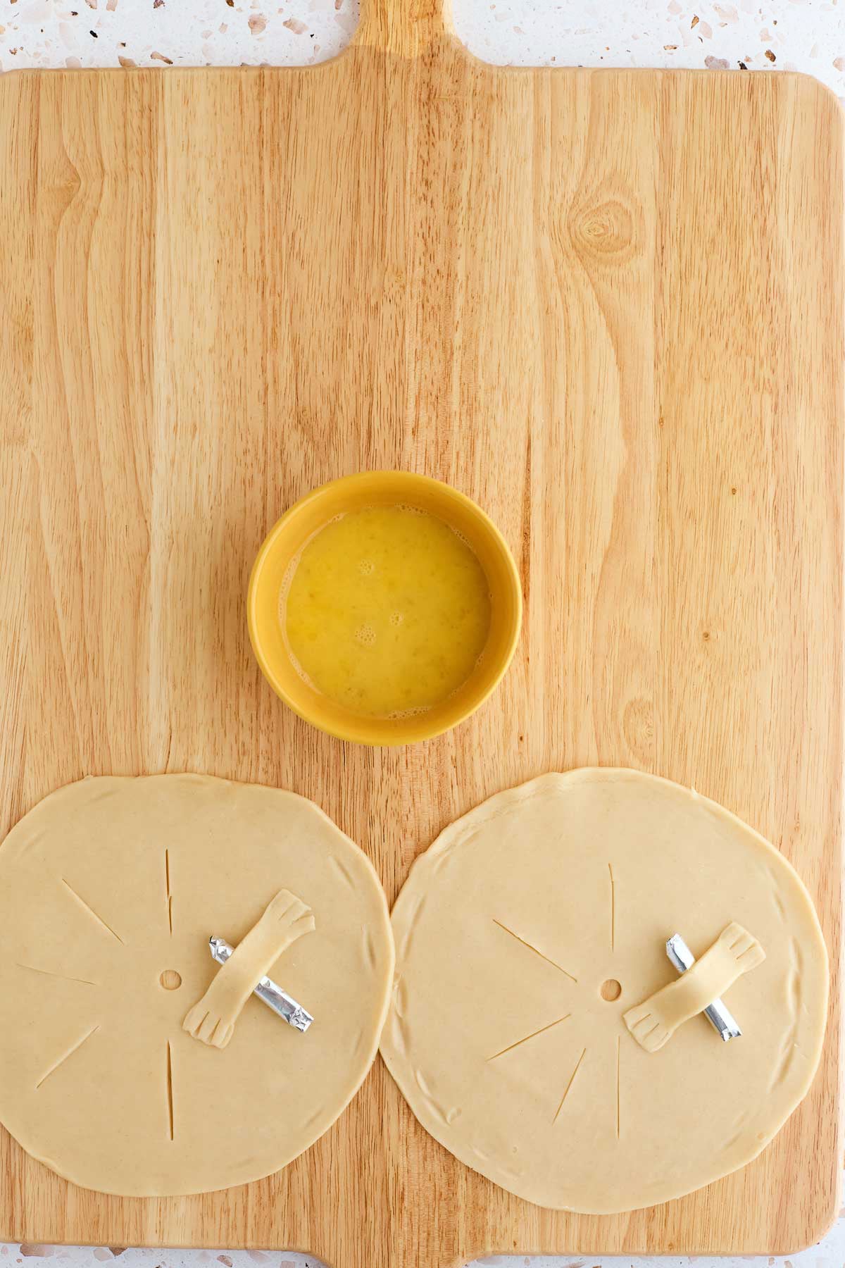 Two uncooked pastry circles with vent slits and foil-wrapped centers on a wooden board, alongside a bowl of beaten egg.