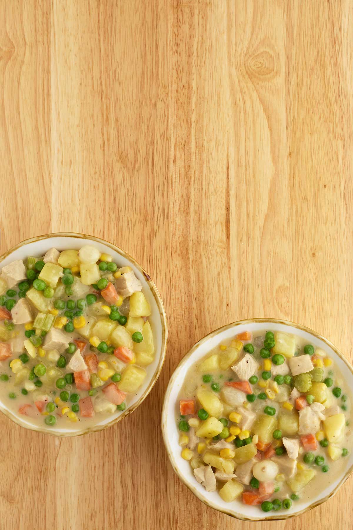Two bowls of chicken pot pie filling with diced chicken, peas, carrots, corn, and potatoes on a wooden surface.