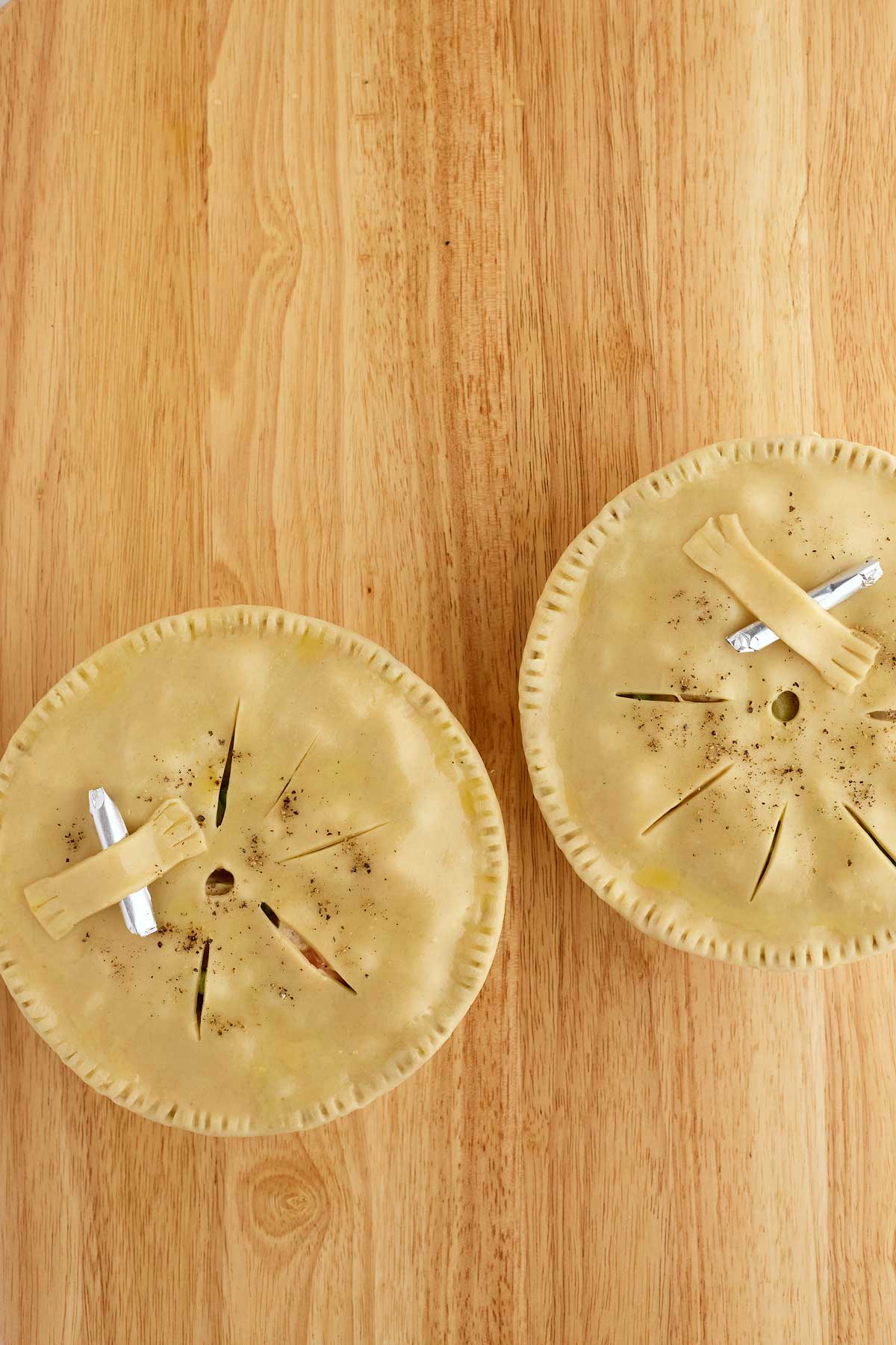 Two uncooked pies with slit tops and strips of dough on a wooden surface.