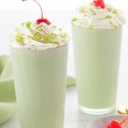 Two green milkshakes topped with whipped cream and cherries, garnished with green sprinkles. A dish of cherries and gold and green coins are nearby.