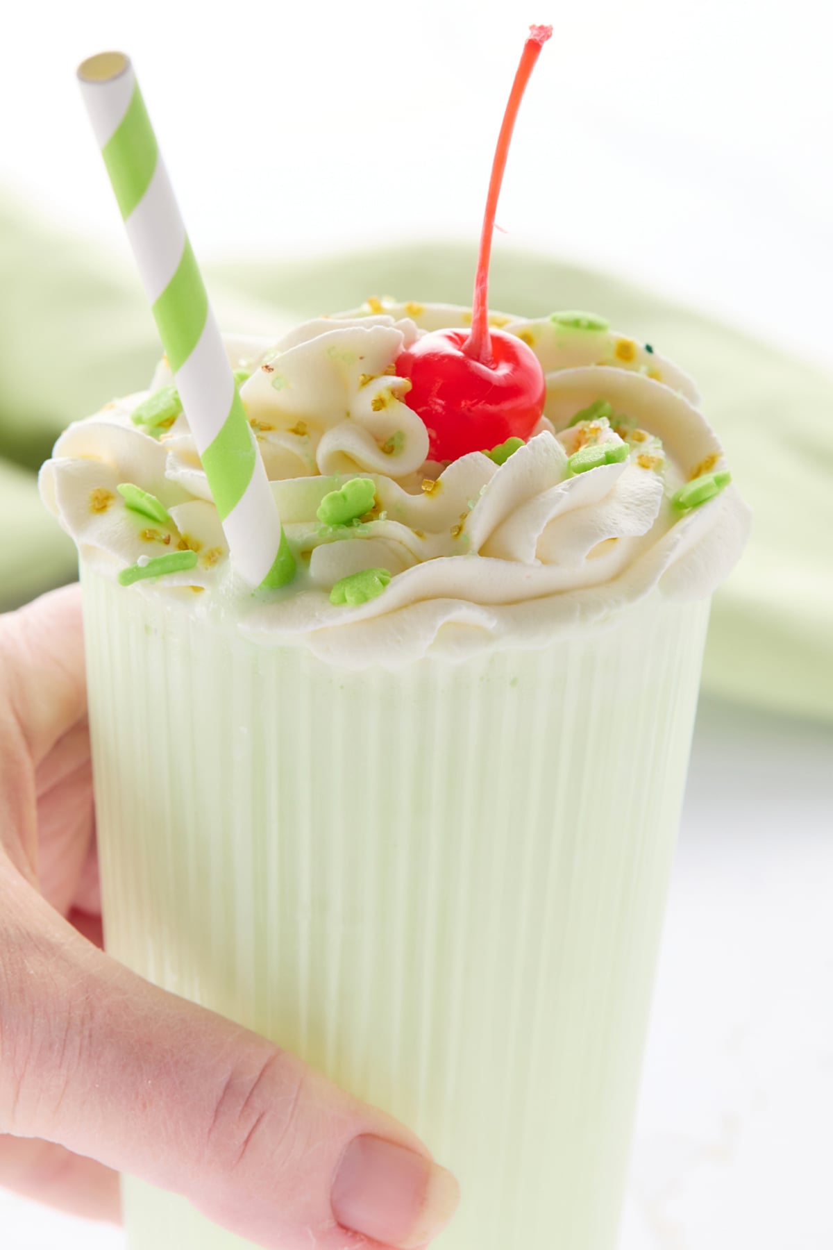 A hand holds a green milkshake, reminiscent of the iconic McDonald's Shamrock Shake, topped with whipped cream, green sprinkles, and a cherry. A green and white striped straw is inserted in the drink.
