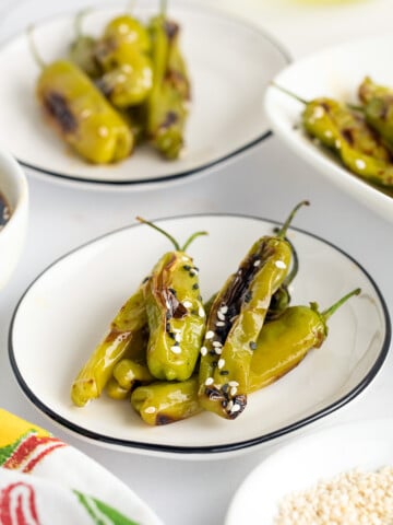 Plates of grilled green shishito peppers, sprinkled with sesame seeds, on a white table.
