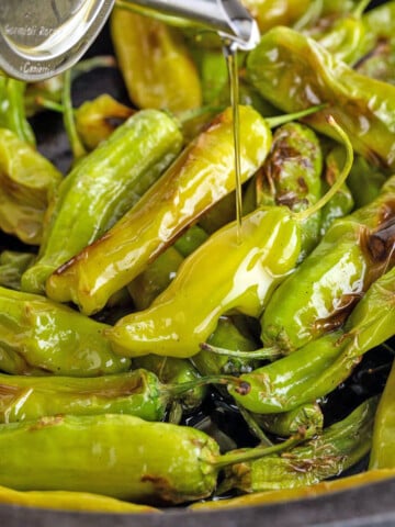 Olive oil is being poured over a pan of blistered green shishito peppers.