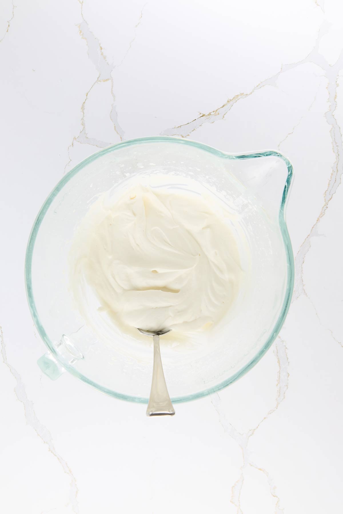 A glass bowl with smooth, white whipped cream and a spoon on a light marble surface.