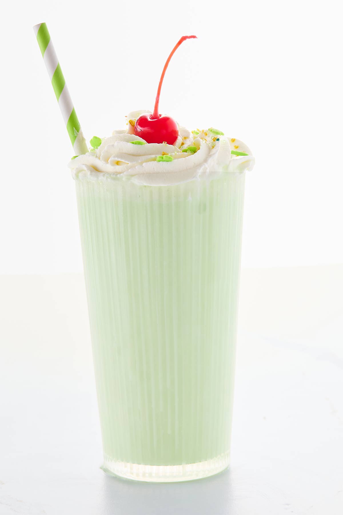 A tall glass of green milkshake topped with whipped cream, a cherry, and sprinkles, with a green and white striped straw.