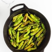 Blistered shishito peppers in a cast iron skillet on a white marble surface.