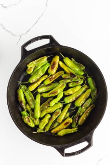 Blistered shishito peppers in a cast iron skillet on a white marble surface.