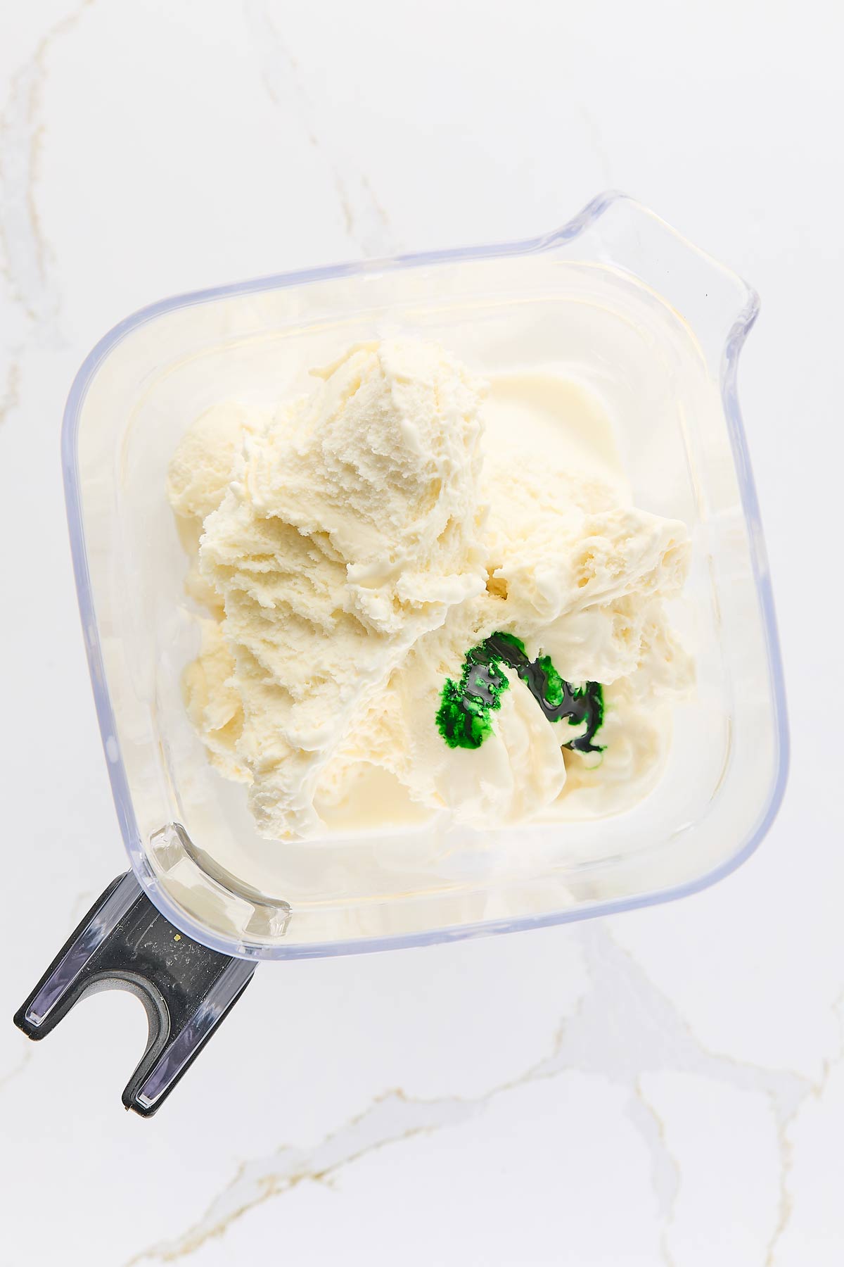 Vanilla ice cream with a drop of green food coloring in a blender container on a white marble surface.