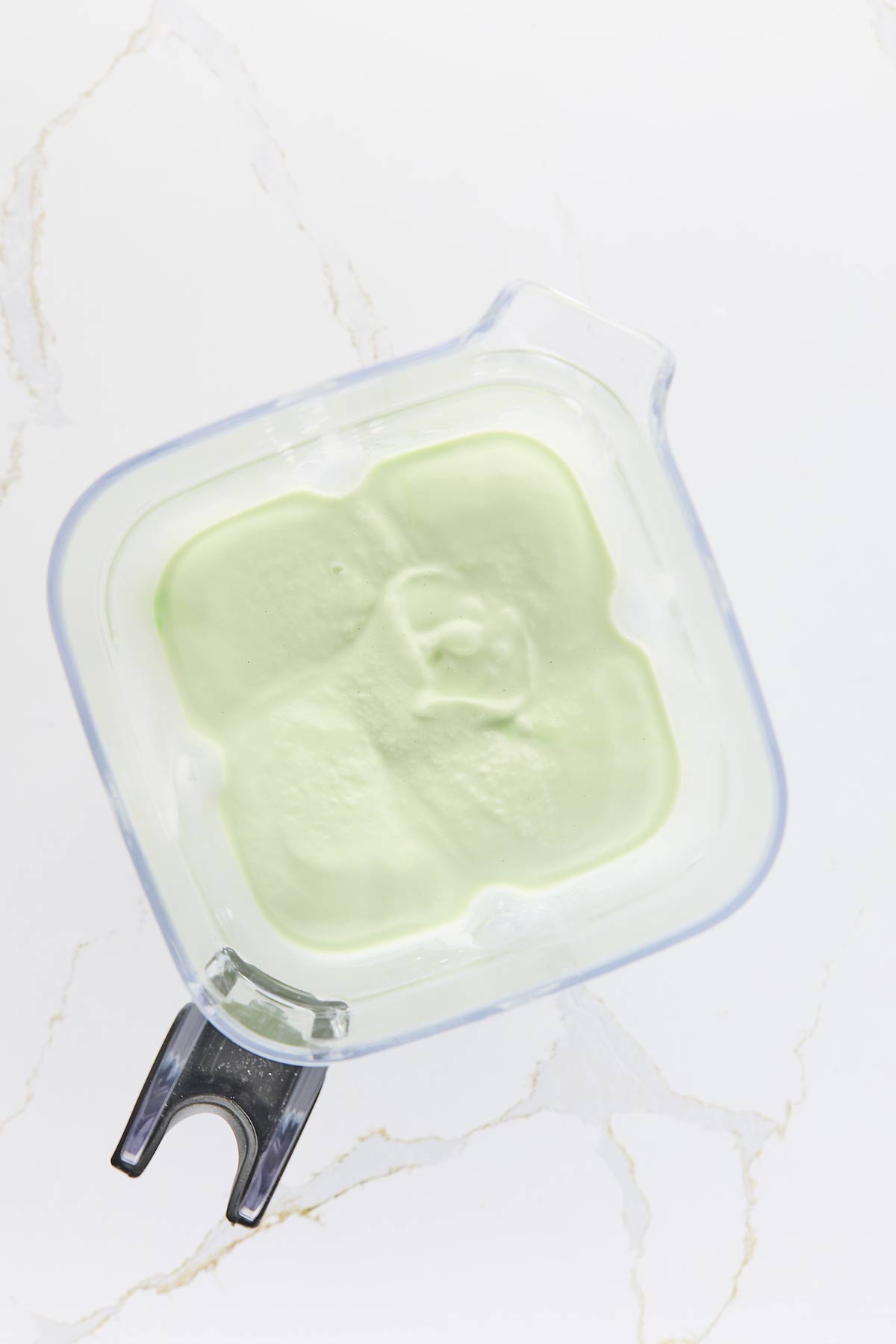 Top view of a blender with light green milkshake inside, placed on a white marble surface.
