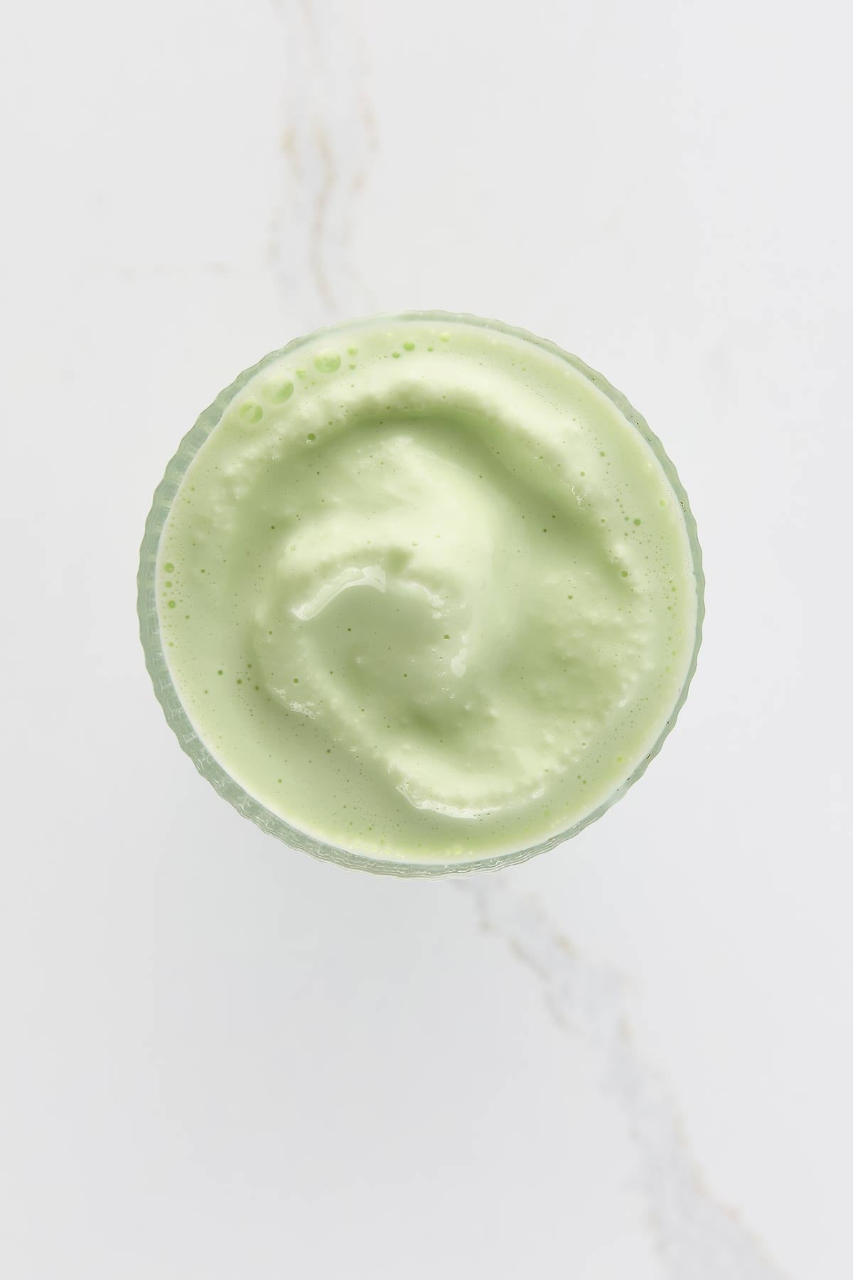 Top view of a frothy, light green milkshake in a glass on a white marble surface.