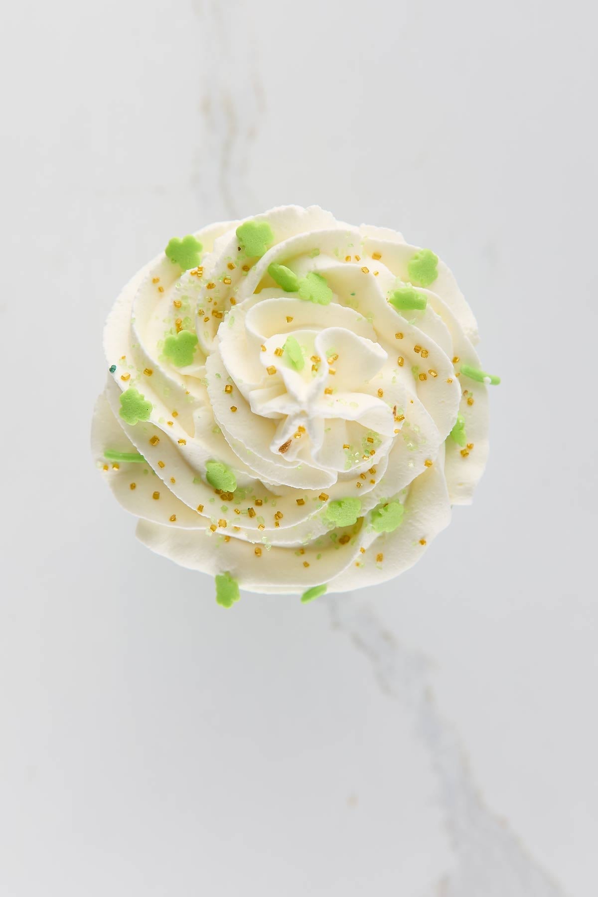 Whipped cream, topped with green clover-shaped sprinkles and yellow sugar pearls, set against a white background.