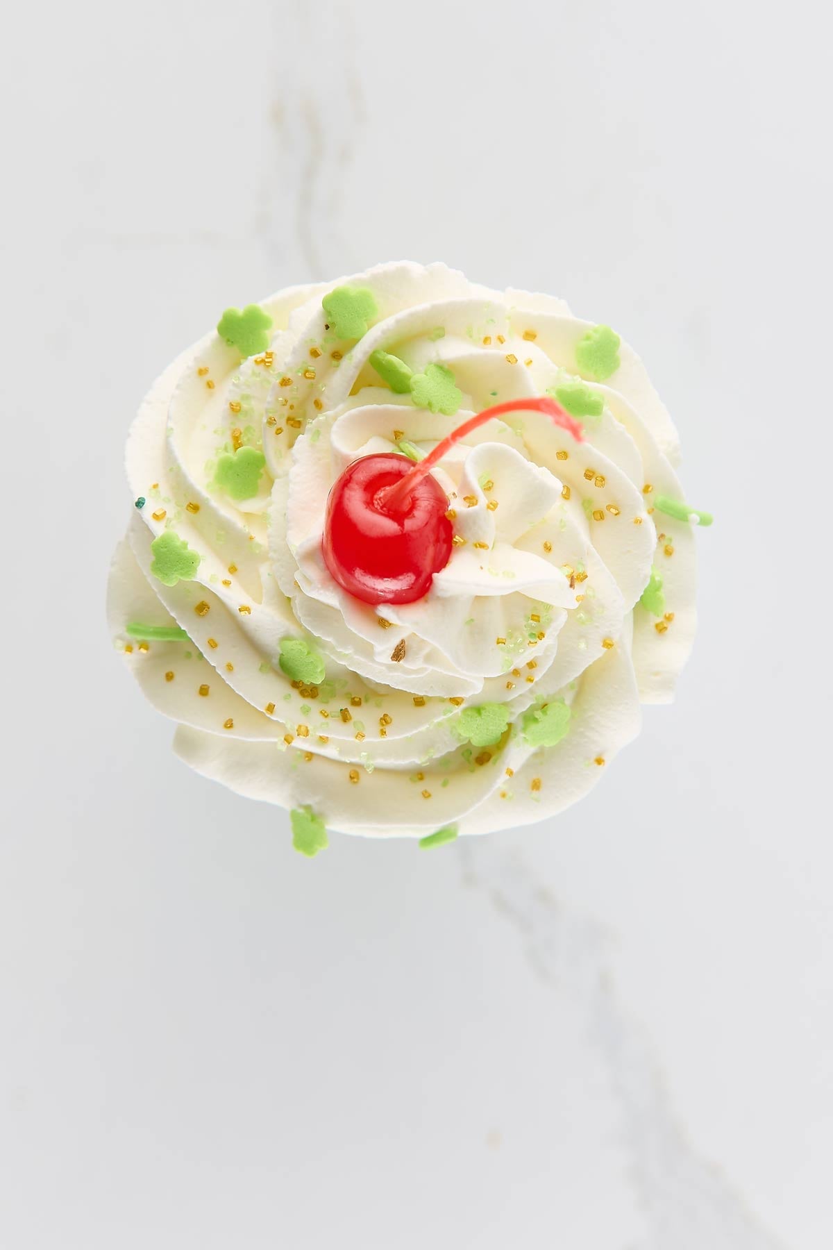 A close-up of awhipped cream with green sprinkles, yellow sugar pearls, and a red cherry on top, viewed from above.