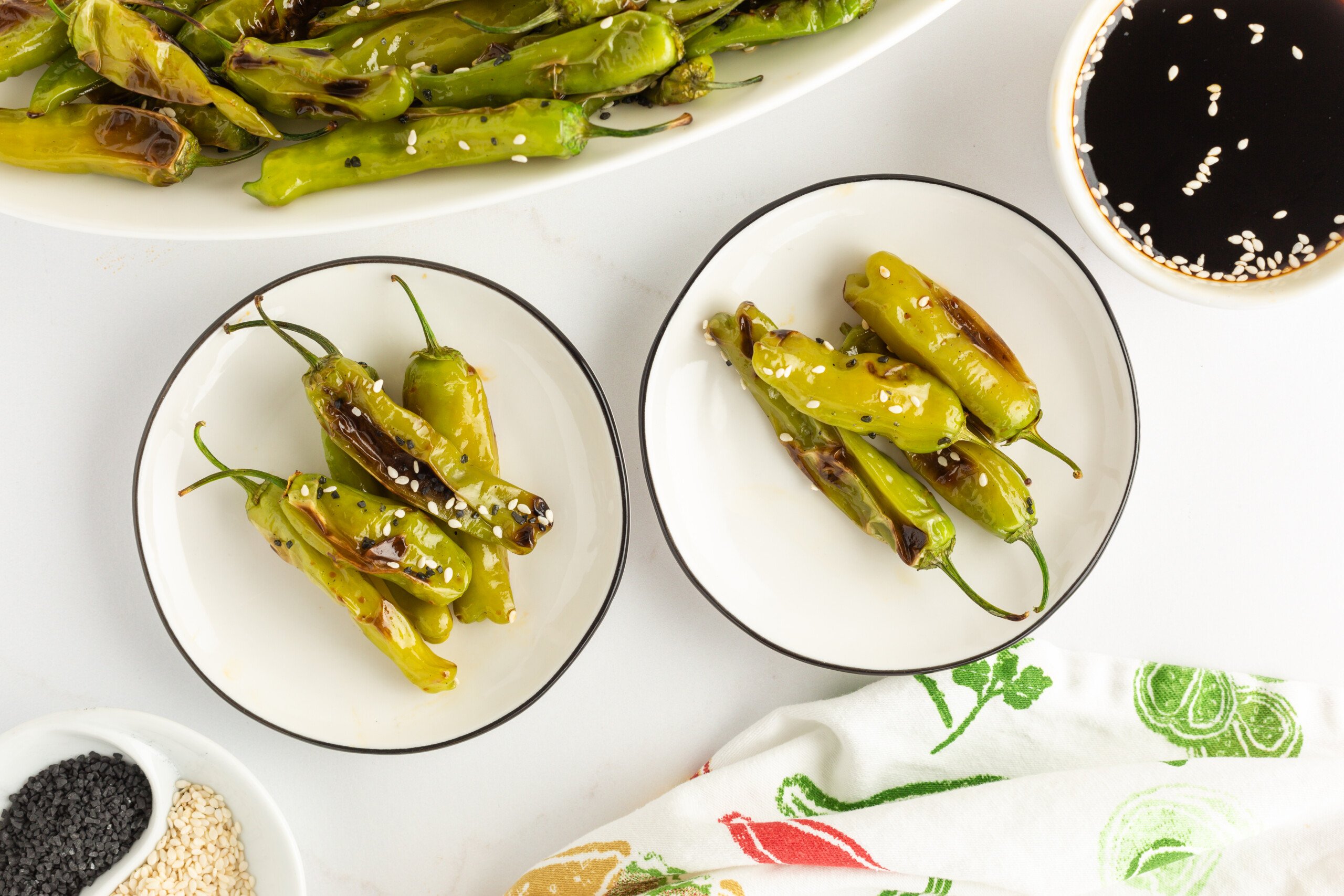 Two small plates with charred shishito peppers sprinkled with sesame seeds, next to a bowl of soy sauce and a larger dish of more peppers on a white surface.