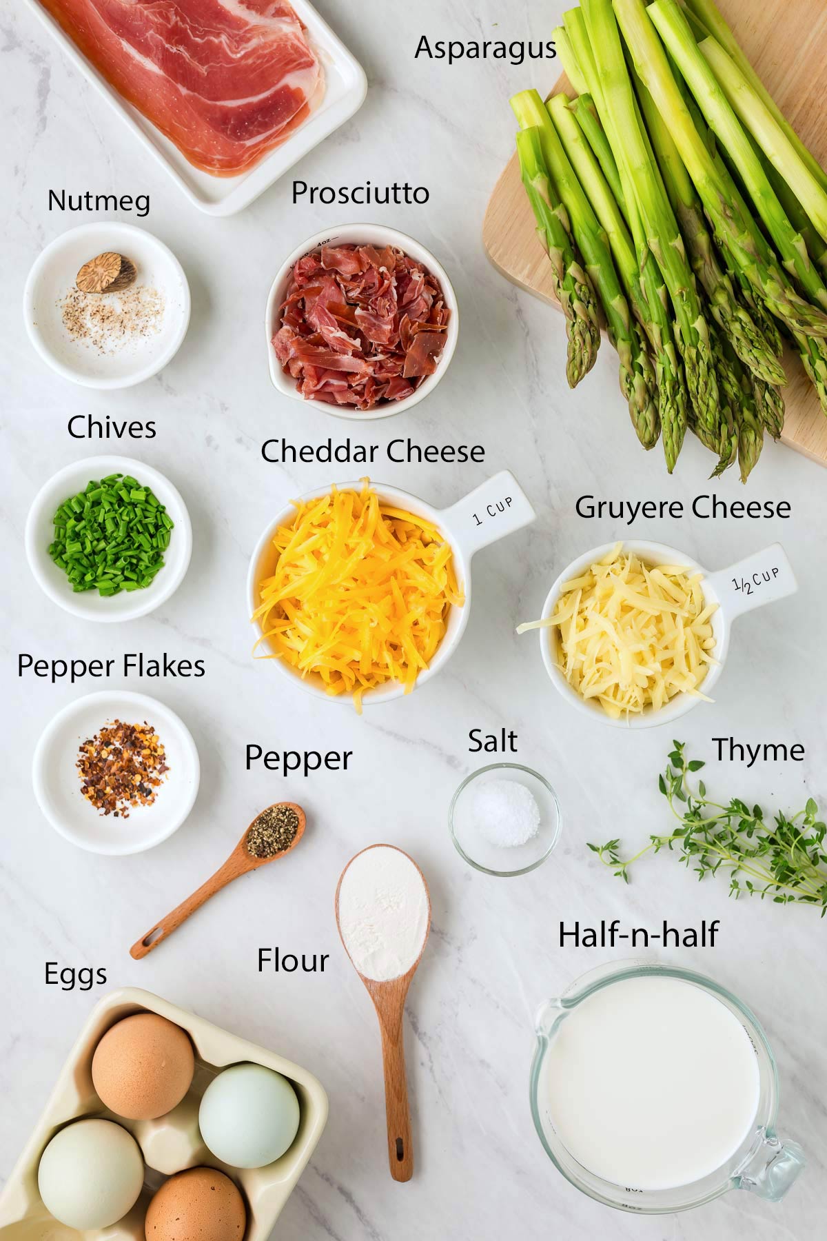 Ingredients for an asparagus quiche, including asparagus, prosciutto, cheddar, gruyere, eggs, half-n-half, flour, pepper, salt, pepper flakes, chives, nutmeg, and thyme on a countertop.