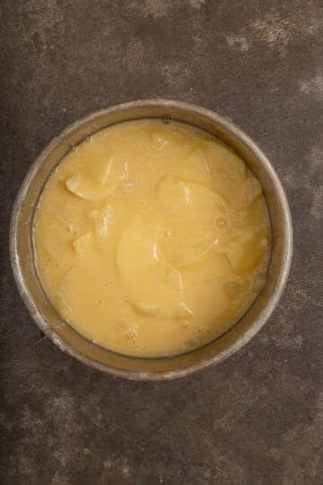 A round metal pan filled with sliced apples coated in yellow pie filling sits on a dark, textured surface.