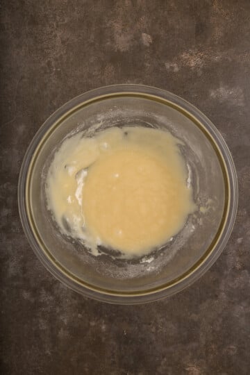 A glass bowl containing a light yellow batter or mixture sits on a dark, textured surface.