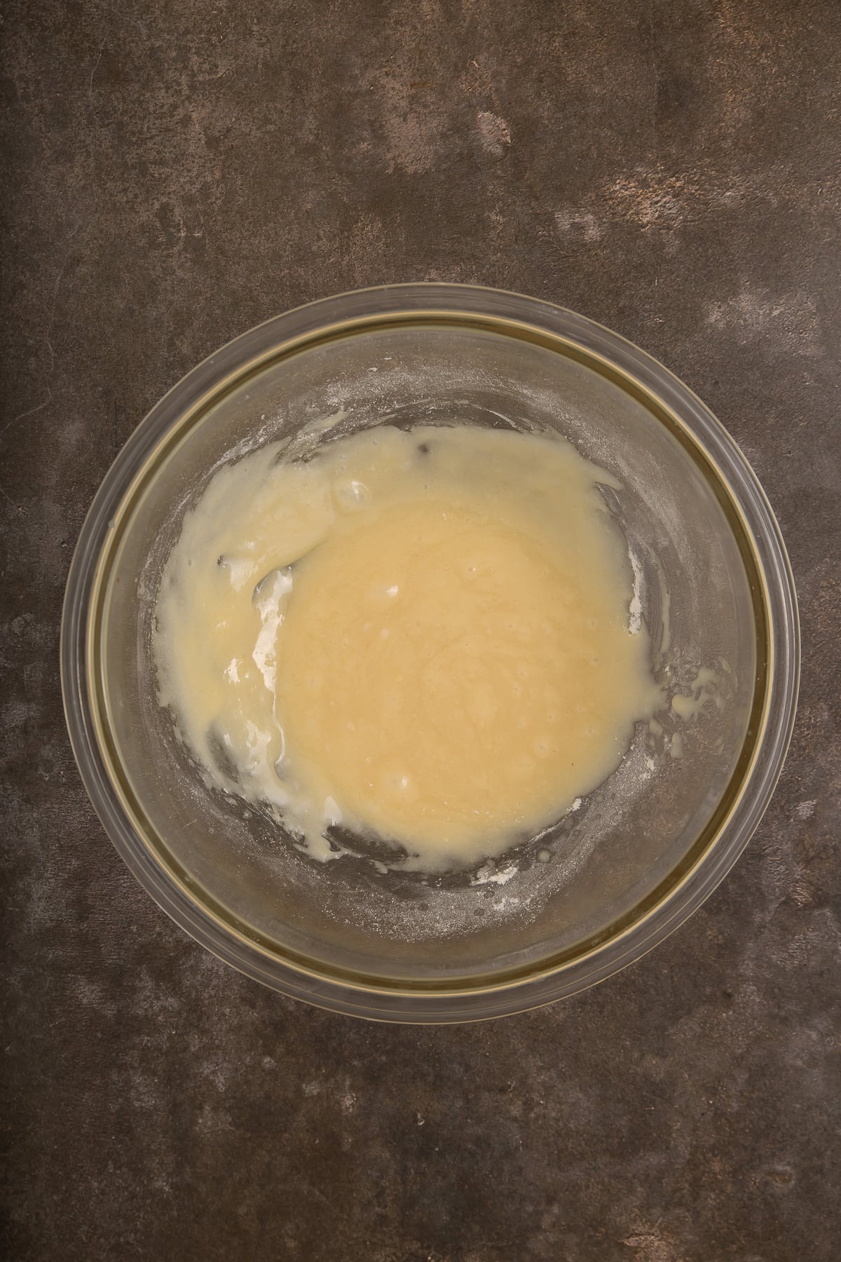 A glass bowl containing a light yellow batter or mixture sits on a dark, textured surface.