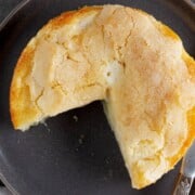 A round, golden-brown pie with a cracked crust, missing one slice, sits on a dark plate.