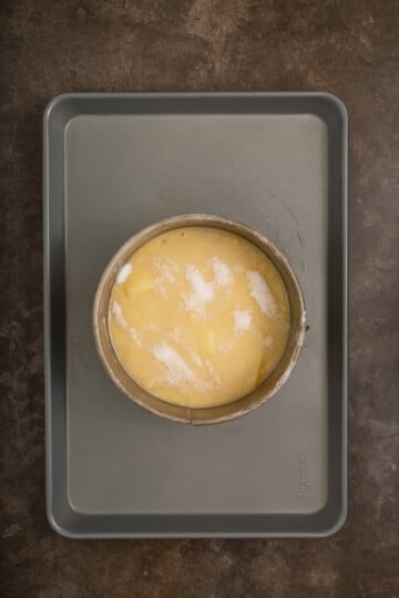 A round cake pan with raw yellow batter topped with sugar sits on a grey baking sheet against a dark countertop background.