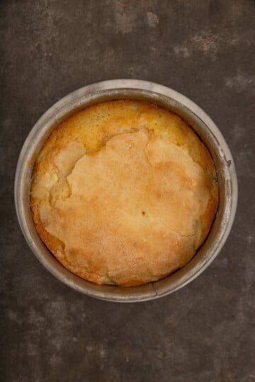 A round, golden-brown cake in a metal baking pan, shown from above, set on a dark, textured surface.