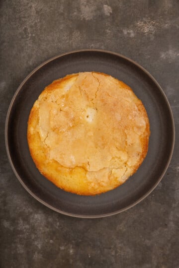 A round, golden-brown cake with a cracked top sits on a dark plate against a textured, dark surface background.