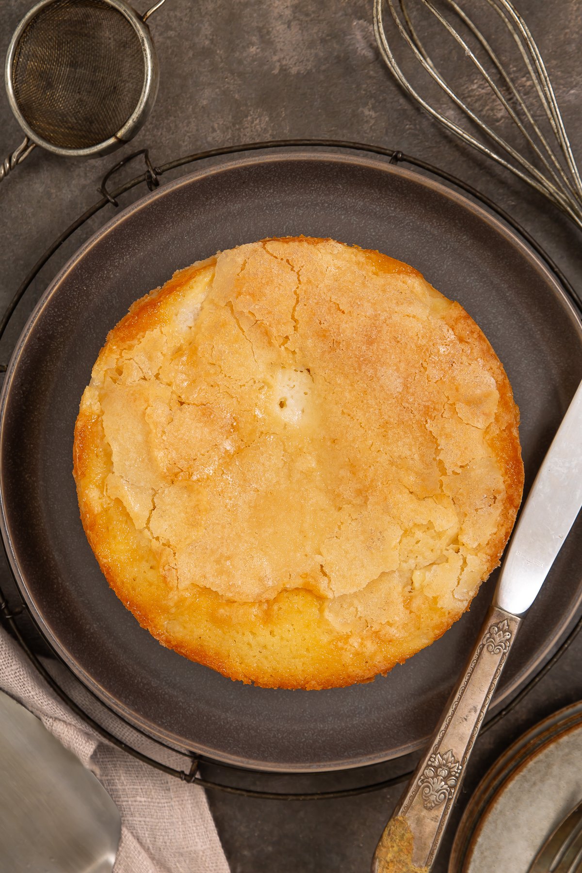 A round, golden-brown French Apple Cake with a cracked top sits on a dark plate, surrounded by a knife, whisk, and metal strainer—inviting you to try this classic recipe French apple cake.