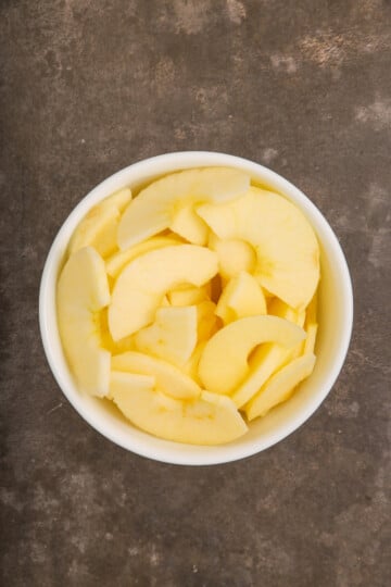 A white bowl filled with peeled and sliced apples, placed on a brown textured surface.
