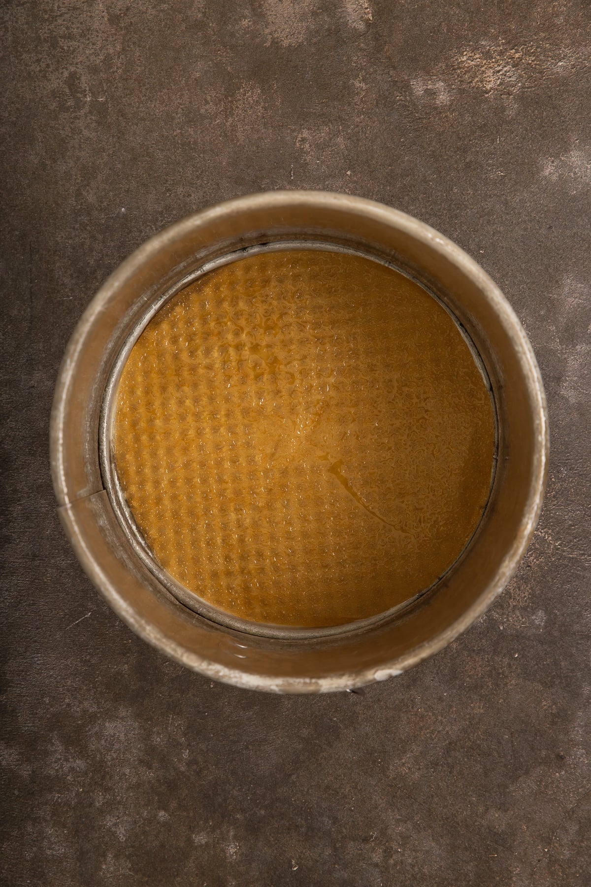A round metal cake pan with an unbaked, textured yellow crust or batter inside, placed on a dark, worn surface.