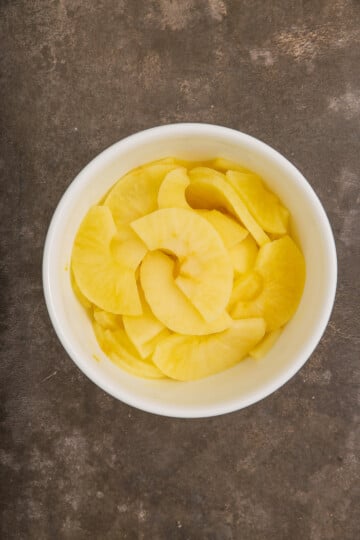 A white bowl filled with thinly sliced, yellow pickled radish sits on a dark textured surface.