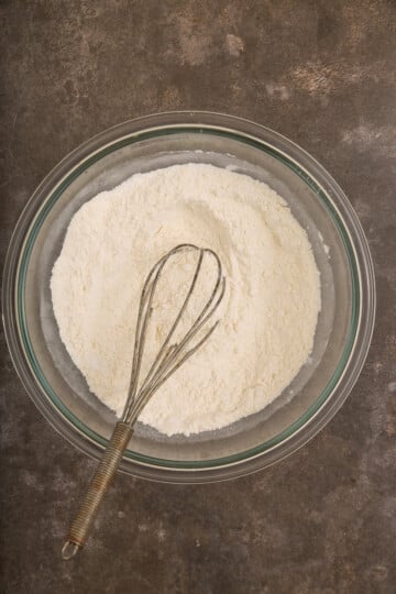 A glass bowl filled with flour mixture and a metal whisk on a dark countertop.