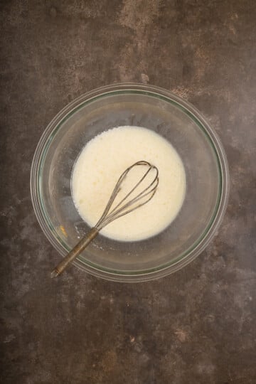 A glass bowl containing a pale liquid mixture with a metal whisk resting inside, placed on a dark textured surface.