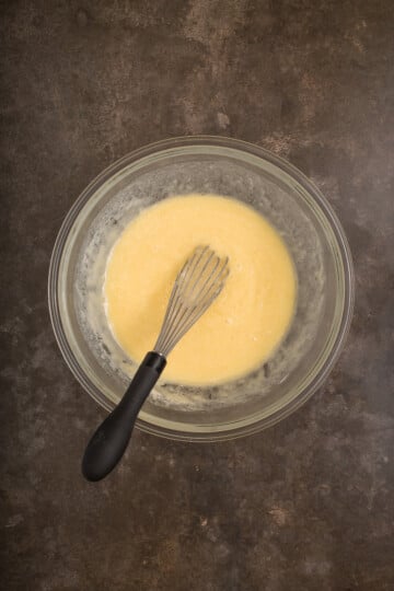 A glass bowl filled with yellow batter and a metal whisk with a black handle resting inside, placed on a dark countertop.