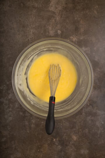 A glass bowl with a yellow batter being mixed by a metal whisk on a dark countertop.