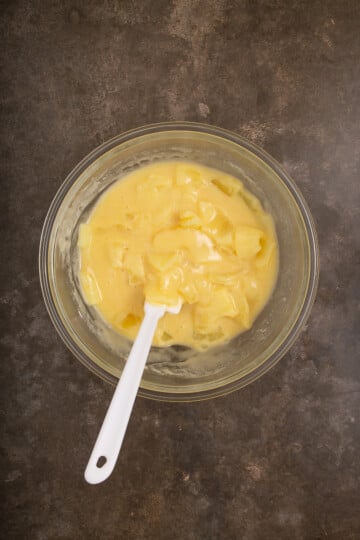 A glass bowl of yellow custard is being mixed with a white spatula on a dark countertop.
