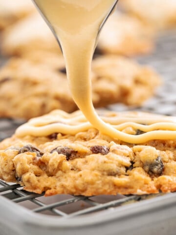 A close-up of icing being drizzled onto an oatmeal raisin cookie on a wire rack, capturing the finishing touch for the best oatmeal raisin cookies recipe.