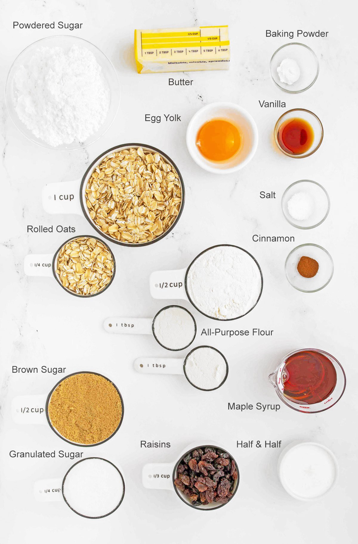 Overhead view of various labeled baking ingredients in bowls, including butter, egg yolk, sugars, oats, flour, raisins, maple syrup, and spices, arranged on a white surface.