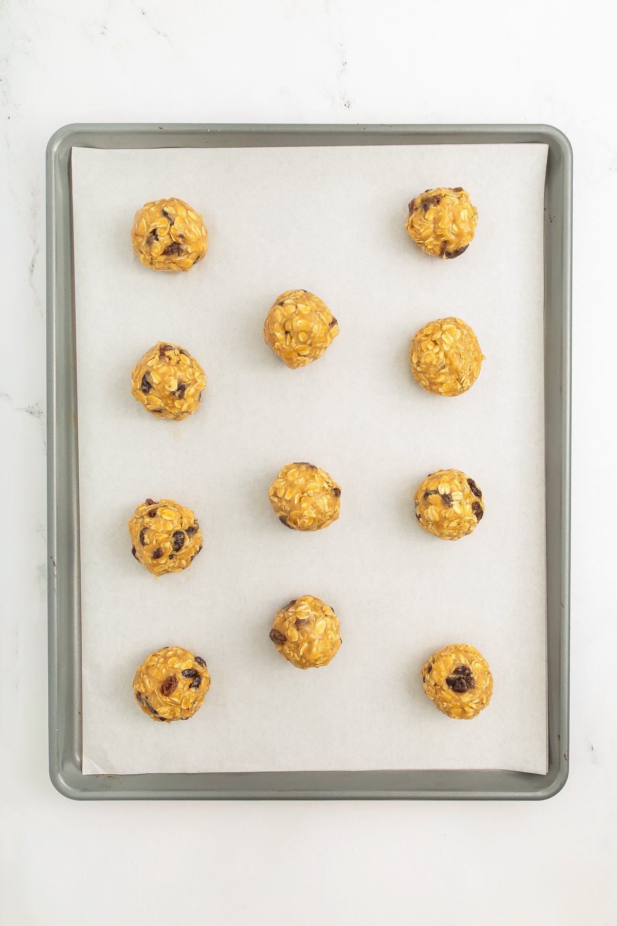A baking sheet lined with parchment paper holds eleven evenly spaced balls of cookie dough with oats and chocolate chips.