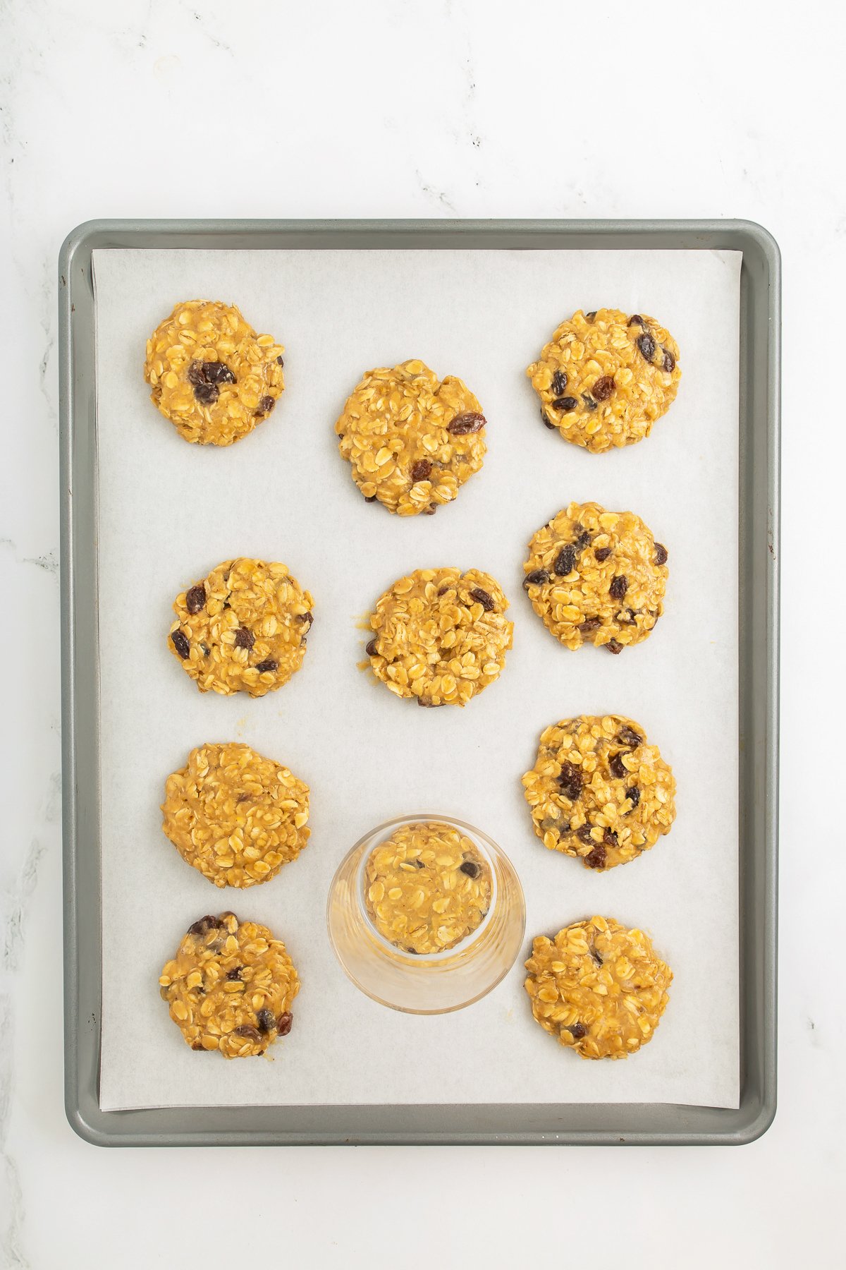A baking tray with eleven unbaked oatmeal raisin cookies on parchment paper, with a glass jar being used to flatten one of the cookies.