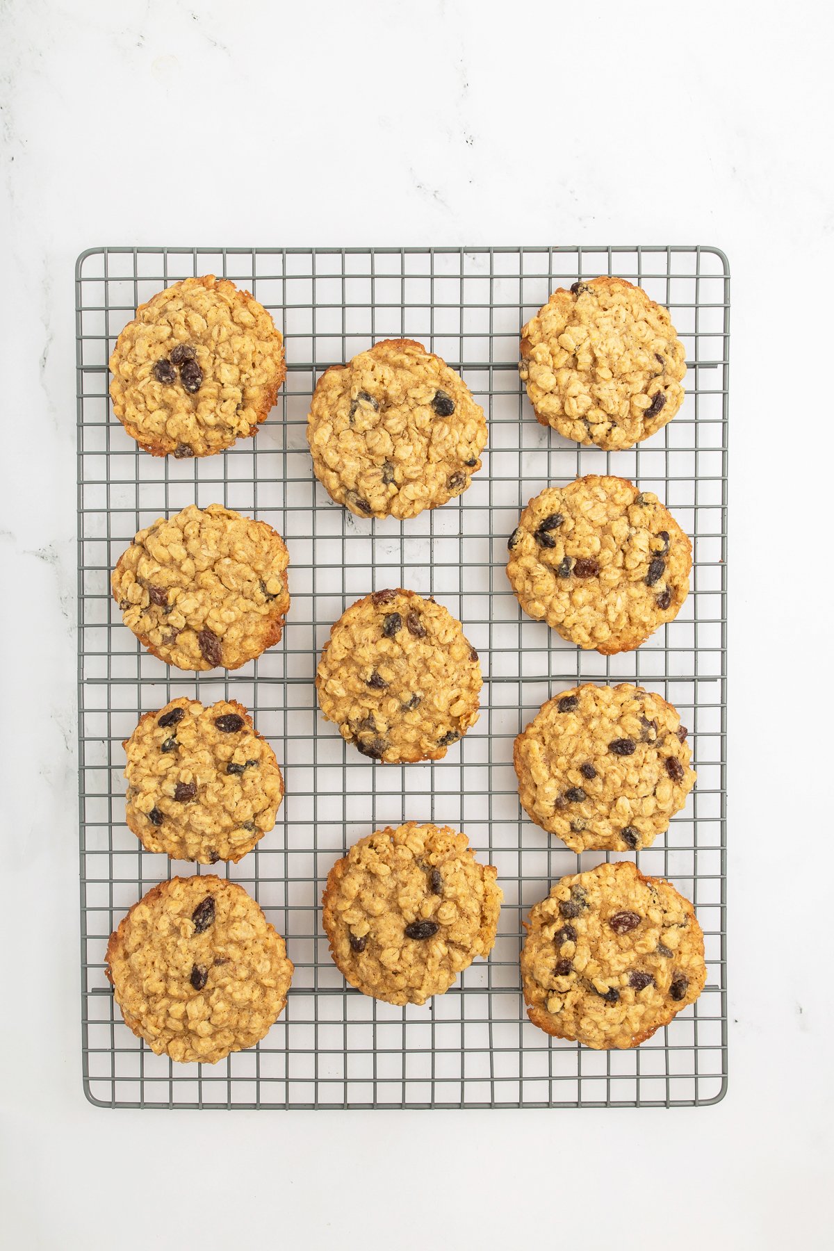 Twelve oatmeal raisin cookies are arranged in rows on a cooling rack set on a white surface.