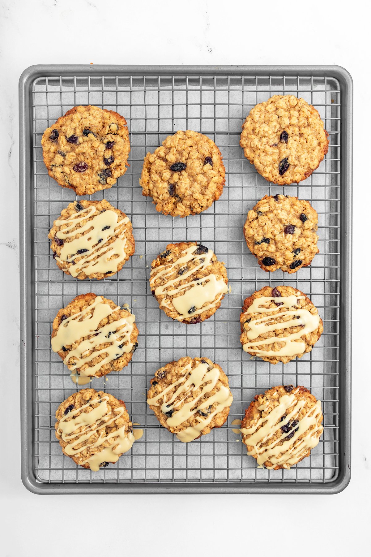 A baking tray with a cooling rack holds twelve oatmeal cookies, some topped with white icing drizzle and others plain with visible raisins.