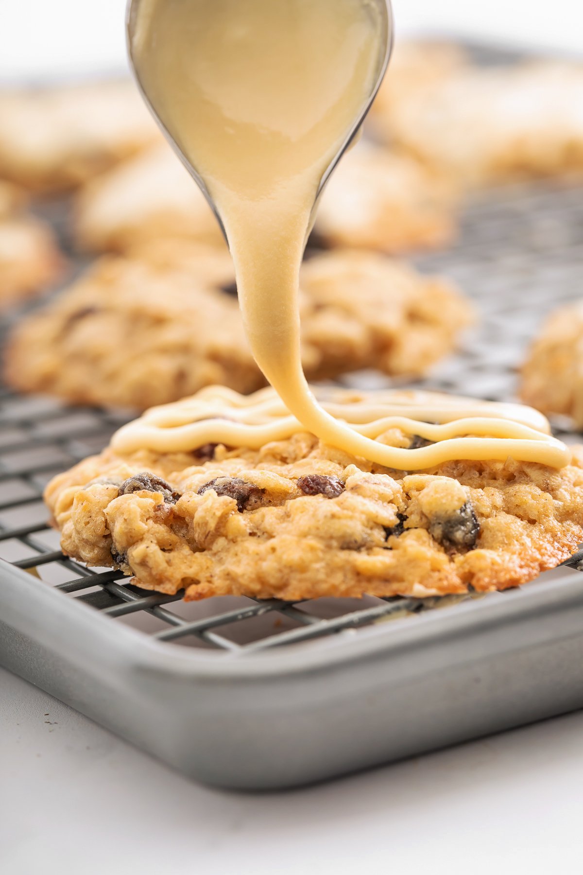 Icing is being drizzled onto an oatmeal raisin cookie on a cooling rack, with several other cookies in the background.