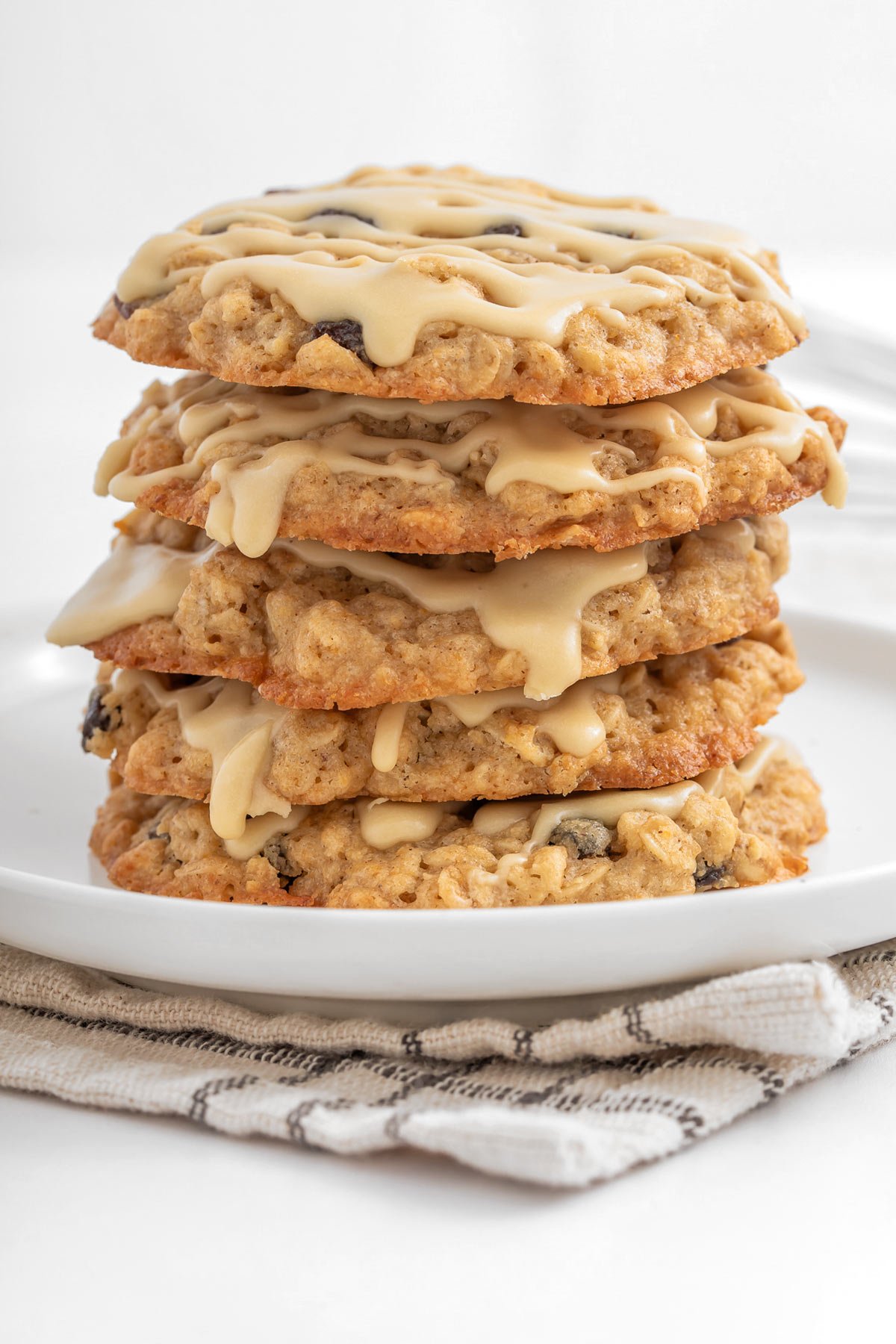 A stack of five oatmeal cookies with icing drizzled on top, placed on a white plate over a folded cloth napkin.