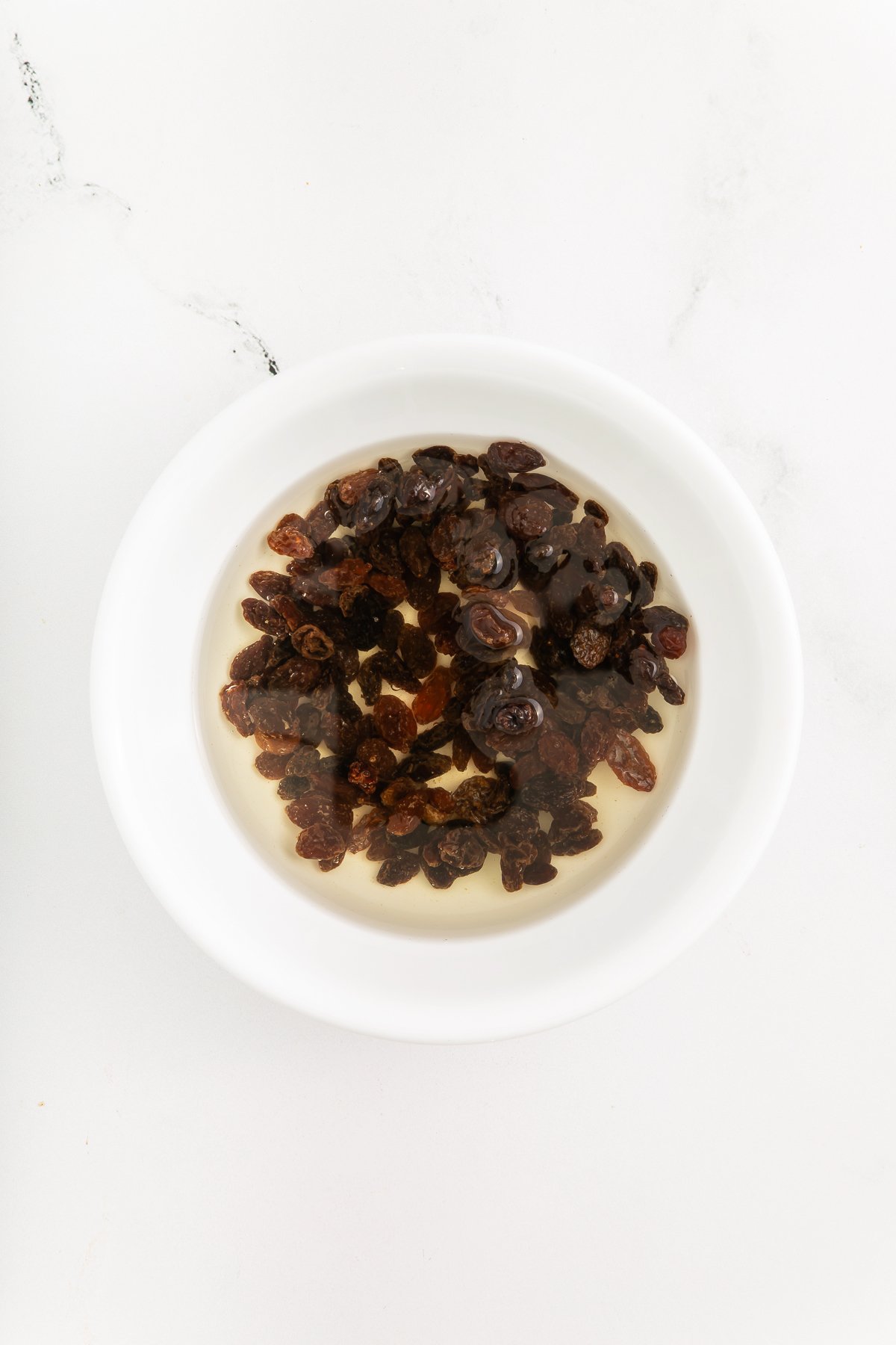 A white bowl filled with raisins soaking in water on a white marble surface.